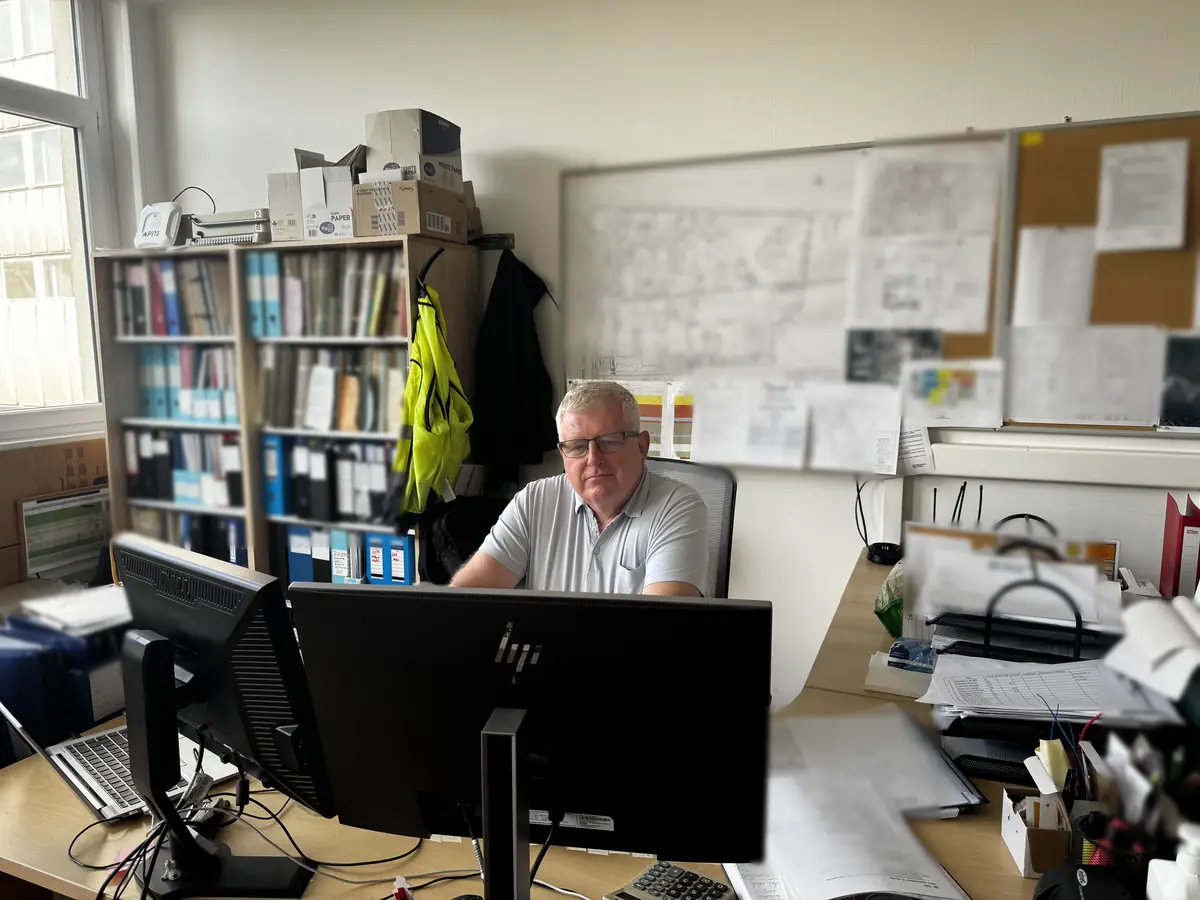 Munkatárs íróasztalnál, mögötte a falon tervrajzok